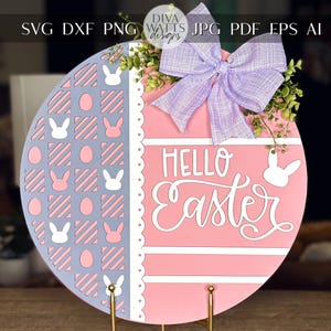 May include: A pink and gray round wooden sign with a scalloped edge. The sign has a white bunny rabbit pattern on the left side and the words "Hello Easter" on the right side. The sign is decorated with a purple bow.