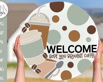 Könnte beinhalten: Ein rundes Willkommensschild mit einem braunen, weißen und grünen Kaffeetassen-Design. Das Schild hat einen weißen Hintergrund mit braunen und grünen Punkten. Der Text auf dem Schild lautet "WELCOME HOPE YOU BROUGHT COFFEE".
