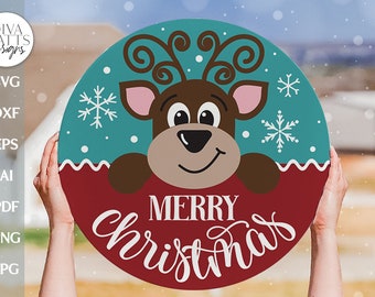 May include: A round wooden sign with a red background and a teal border. A cartoon reindeer with brown fur, pink ears, and a brown nose peeks over the top of the sign. The sign says "Merry Christmas" in white cursive lettering.