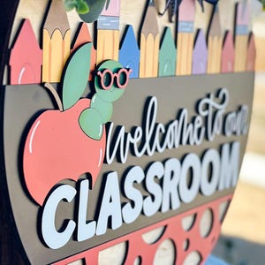 Puede incluir: Un letrero de bienvenida de madera con una manzana roja, un gusano verde con gafas y el texto "Bienvenido a nuestra aula".