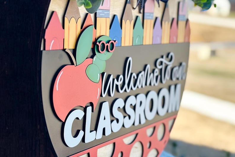 Puede incluir: Un letrero de bienvenida de madera con una manzana roja, un gusano verde con gafas y el texto "Bienvenido a nuestra aula".