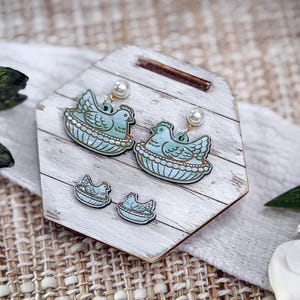 May include: A set of earrings and stud earrings featuring a blue bird in a nest design. The earrings have a pearl accent. The jewelry is displayed on a white wooden stand with a rustic aesthetic.