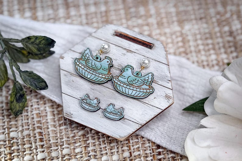 May include: A set of earrings and stud earrings featuring a blue bird in a nest design. The earrings have a pearl accent. The jewelry is displayed on a white wooden stand with a rustic aesthetic.