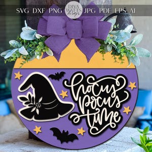 Puede incluir: Un letrero redondo de madera con temática de Halloween, con una combinación de colores púrpura y amarillo. El letrero presenta un sombrero de bruja, murciélagos, estrellas y la frase "Hocus Pocus Time" en escritura blanca. Un lazo morado y vegetación adornan la parte superior.