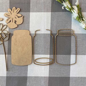 May include: A set of three wooden craft cutouts, including two mason jar shapes and a flower with leaves. The cutouts are on a white and black checkered background.