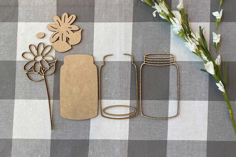 May include: A set of three wooden craft cutouts, including two mason jar shapes and a flower with leaves. The cutouts are on a white and black checkered background.