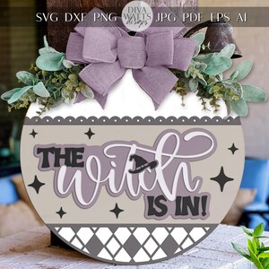 May include: A round wooden sign with the text "THE WITCH IS IN!" in a layered design. The sign features a purple bow, greenery, and star accents. A witch hat is incorporated into the design. The sign is suitable for Halloween.