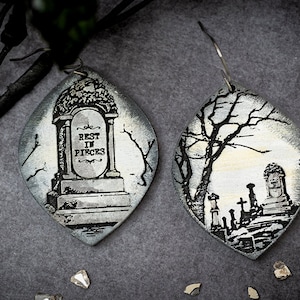 May include: Oval-shaped earrings with a spooky graveyard design. One earring features a tombstone with "REST IN PIECES" and the other shows a cemetery scene with bare trees. The earrings are silver and black.
