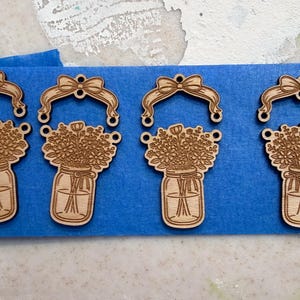 May include: Four laser-cut wooden ornaments. Each ornament features a mason jar filled with flowers, topped with a bow. The ornaments are light brown and are arranged on a blue surface.
