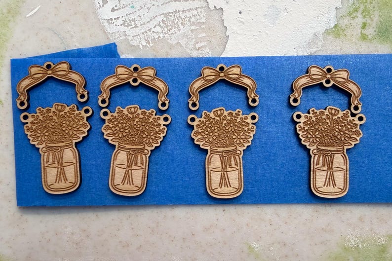 May include: Four laser-cut wooden ornaments. Each ornament features a mason jar filled with flowers, topped with a bow. The ornaments are light brown and are arranged on a blue surface.