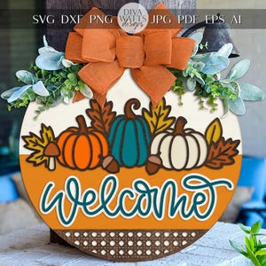 May include: A round wooden welcome sign with a fall theme. It features orange, teal, and white pumpkins, fall leaves, and the word "Welcome" in teal script. The sign is decorated with a large orange bow and greenery.