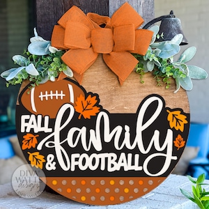 Op de afbeelding: Een ronde houten plank met de woorden "Fall, Family & Football" in witte letters en een voetbalafbeelding. Het heeft een oranje strik, groen en herfstbladeren. Een decoratieve bel is bevestigd.