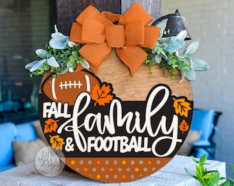 May include: A round wooden sign with the words "Fall, Family & Football" in white script and a football graphic. It features an orange bow, greenery, and fall leaves. A decorative bell is attached.