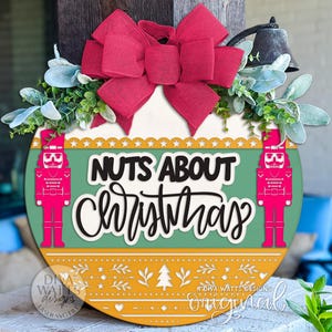 May include: A round Christmas sign with the text "Nuts About Christmas" in black and white script. The sign features two pink nutcracker figures, a red bow, and greenery. The bottom has a yellow and orange pattern.