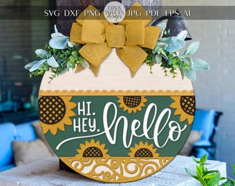 Può includere: Un cartello di benvenuto rotondo in legno con un design a girasole e fiocco. Il cartello presenta le parole "Hi. Hey. hello" in scrittura bianca su sfondo verde, con accenti di girasole. Il cartello è decorato con verde e un fiocco giallo.