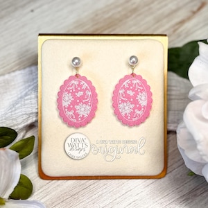 May include: A pair of pink oval earrings with a white floral design and a pearl accent. The earrings are displayed in a gold-trimmed box with the text "DIVA WATTS DESIGNS" and "original".