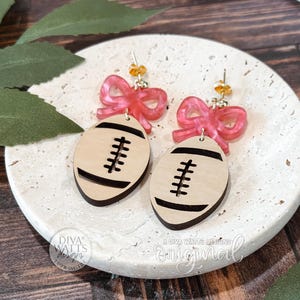 Puede incluir: Pendientes con dise&ntilde;o de bal&oacute;n de f&uacute;tbol. Los pendientes tienen forma de bal&oacute;n de f&uacute;tbol de color madera clara con detalles negros, combinados con lazos rosas. Los pendientes se exhiben sobre una superficie blanca texturizada.