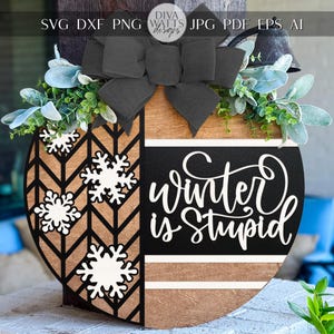 May include: Wooden winter door decoration with a black bow and foliage. The design features a chevron pattern, snowflakes, and the phrase "Winter is Stupid" in white script.