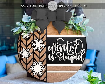 May include: Wooden winter-themed door hanger with a black bow and greenery. The design includes a chevron pattern, snowflakes, and the phrase "Winter is Stupid" in white script.