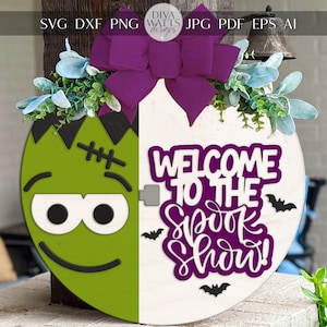 May include: A Halloween door decoration. One half shows a green Frankenstein face, the other half has the text "WELCOME TO THE SPOOK SHOW!" in purple. The design is decorated with a purple bow and artificial greenery.