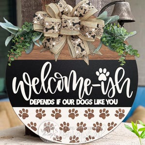 May include: A round wooden welcome sign with a black and brown color scheme. The sign features the text "Welcome-ish" and "Depends if our dogs like you" in white script. The bottom half has paw prints. A bow with dog bone accents and greenery adorns the top.