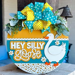 May include: A round wooden door hanger with a blue and yellow polka dot bow, green leaves, and a white goose with the text "Hey Silly Goose" in white letters.