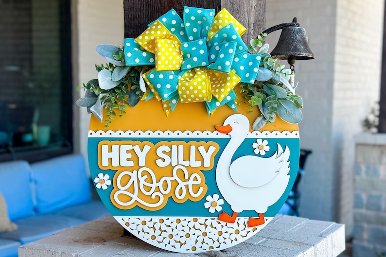 May include: A round wooden door hanger with a blue and yellow polka dot bow, green leaves, and a white goose with the text "Hey Silly Goose" in white letters.