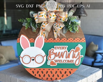 Pode incluir: Um sinal redondo de madeira com fundo verde e um coelho branco com óculos. O sinal diz "Every Bunny Welcome" com um design de cenoura. O sinal é decorado com um laço marrom com um padrão de cenoura branco e amarelo.