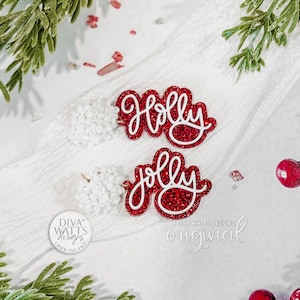 Op de afbeelding: Twee rode glitter kerstversieringen met de woorden "Holly" en "Jolly" in witte letters. De versieringen liggen op een wit oppervlak met takjes groen en rode bessen. Het logo van Diva Watts Designs is zichtbaar.
