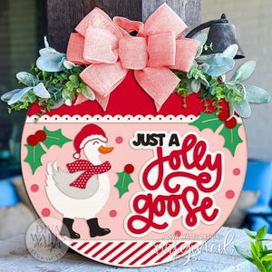 Könnte beinhalten: Ein rundes, rosa-rotes Holzschild mit dem Text "Just a Jolly Goose". Das Schild zeigt eine Cartoon-Gans mit Weihnachtsmütze und Schal. Es ist mit Stechpalmen, einer großen rosa Schleife und Grünzeug verziert.