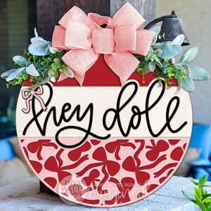 May include: Round wooden door hanger with the words "Hey Doll" in black script. The top half is cream and red, with a red bow and greenery. The bottom half has a red bow pattern. A pink bow and a small bell are at the top.