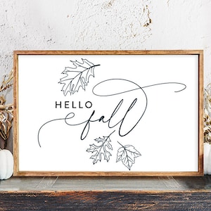 Puede incluir: Un cartel blanco con marco marrón, que presenta el texto "Hello fall" en una fuente cursiva con tres hojas estilizadas. El cartel se muestra sobre una superficie de madera con calabazas y flores secas.