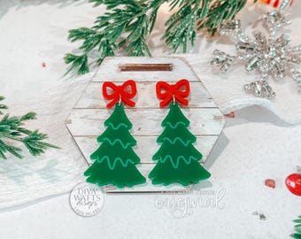 May include: A pair of Christmas tree earrings. Each earring features a green tree shape with a red bow at the top. The earrings are displayed on a white and wood-grain surface, with a snowflake and greenery in the background.