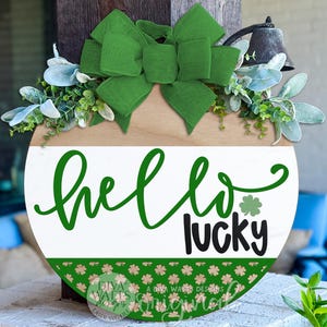 May include: Round wooden St. Patrick's Day sign with the words "hello lucky" in green and black. The sign has a green bow, greenery, and a bell. The bottom of the sign has a green border with shamrock designs.