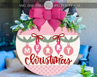 May include: A round, pink Christmas ornament with the words "MERRY Christmas" in red script. The ornament has a pink bow at the top, green and white foliage, and hanging pink ornaments. The background is a blurred outdoor scene.