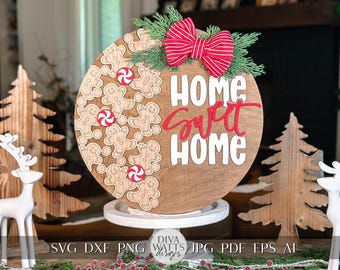 Könnte beinhalten: Ein rundes Holzschild mit den Worten "Home Sweet Home" in weißer und roter Schrift. Das Schild ist mit einer roten und weißen Schleife, grünen Tannenzweigen und Lebkuchenmännern mit Zuckerstangen verziert.