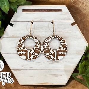 May include: A pair of wooden earrings featuring a brown and white football design. The earrings are round with a central hole and have gold hoops.