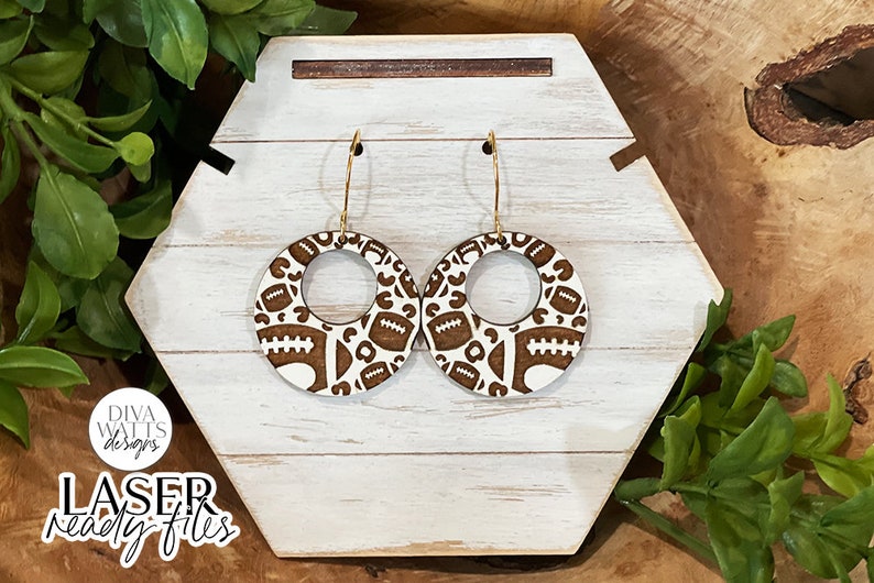 May include: A pair of wooden earrings featuring a brown and white football design. The earrings are round with a central hole and have gold hoops.