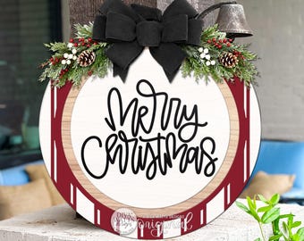 Pode incluir: Uma placa de porta redonda de madeira com o texto Merry Christmas em letra cursiva preta. Possui um centro branco, uma borda com veios de madeira clara e um anel externo vermelho escuro com listras brancas, decorada com um grande laço preto e folhagem.