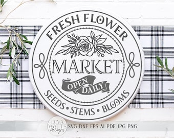 May include: A white round sign with black text that reads "FRESH FLOWER MARKET" with a floral design and a banner that says "OPEN DAILY". The sign also says "SEEDS STEMS BLOOMS".