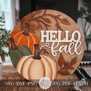 Puede incluir: Un letrero redondo de madera con el texto "Hello Fall" en letras blancas. El letrero está decorado con tres calabazas, dos verdes y una naranja, y hojas marrones.