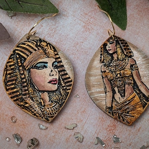 May include: A pair of gold-toned earrings with a laser-cut design of an Egyptian queen. The earrings are made of wood and have a teardrop shape. The queen is wearing a gold headdress and a gold necklace. The earrings are on a light brown background.
