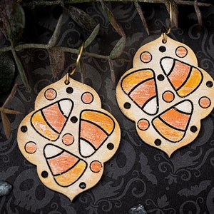 Puede incluir: Un par de pendientes de temática de Halloween con un diseño de maíz dulce blanco, naranja y negro sobre un fondo beige. Los pendientes tienen forma de trébol de cuatro hojas.