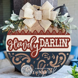 May include: Round wooden sign with the words "Howdy Darlin'" in a decorative font. The sign has a cream, red, and navy blue color scheme with a paisley design. It is decorated with a bow, greenery, and a small bell.