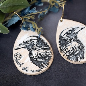 May include: A pair of wooden teardrop-shaped earrings with a black and white raven illustration. One earring has the text "With the raven" and the other earring has the text "Nevermore".