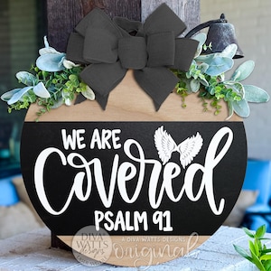 May include: A round wooden sign with a black and natural wood finish. The sign features the words "WE ARE COVERED" in white script, with "PSALM 91" below. It is decorated with a black bow, greenery, and a small bell.
