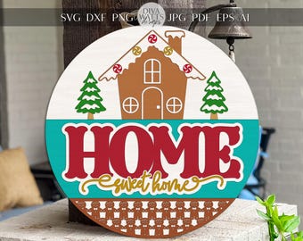 Könnte beinhalten: Runder Holzschild mit Lebkuchenhaus-Design. Das Schild zeigt die Worte "HOME sweet home" in roter und goldener Schrift. Zwei grüne Weihnachtsbäume flankieren das Lebkuchenhaus. Das Schild hat einen Durchmesser von etwa 30 cm.