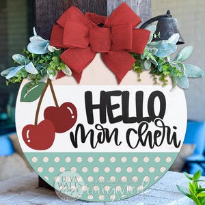 May include: A round wooden sign with a red bow, greenery, and the words "HELLO mon cheri". The sign features two red cherries and a mint green polka dot pattern at the bottom. A decorative item for the home.