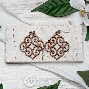 Puede incluir: Un par de pendientes de madera marrón con un intrincado patrón simétrico de volutas en forma de cuatrifolio. Los pendientes están sujetos a postes metálicos de color plateado y se presentan sobre una base rectangular de textura pétrea.