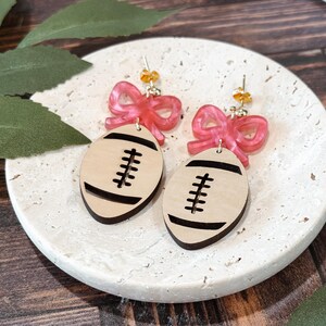 Puede incluir: Pendientes con dise&ntilde;o de bal&oacute;n de f&uacute;tbol. Los pendientes tienen forma de bal&oacute;n de f&uacute;tbol de color madera clara con rayas negras y un lazo rosa. Los pendientes est&aacute;n sobre una superficie blanca texturizada con hojas verdes en el fondo.